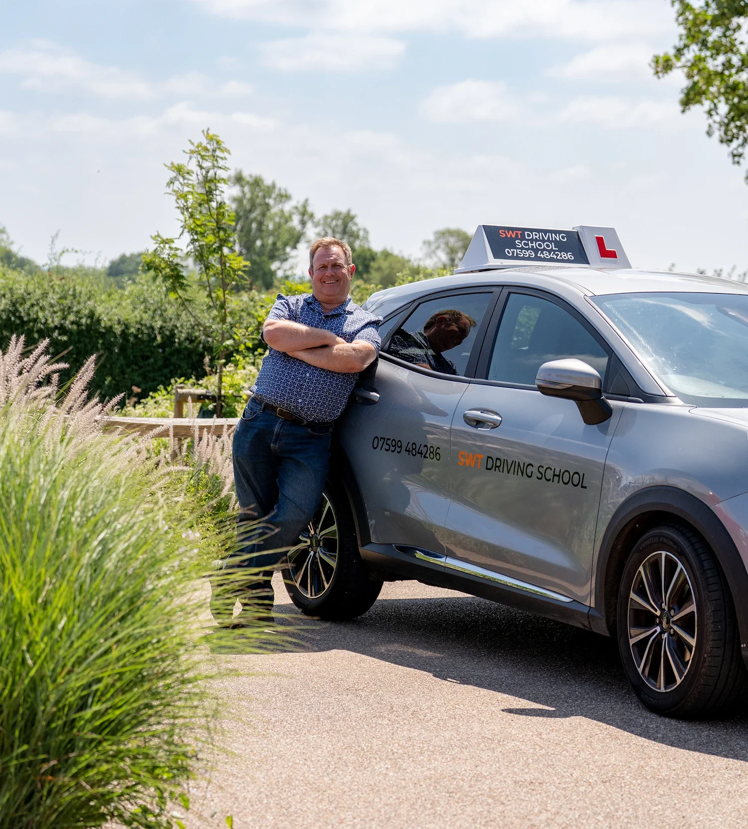 Driving instructor Sean Wyarrt standing next to a driving school car in Westwood Wiltshire, with arms crossed outside on a sunny day. The car is a Ford Puma, and has a rooftop sign with the text 'SWT DRIVING SCHOOL' and a red 'L' learner driver sign