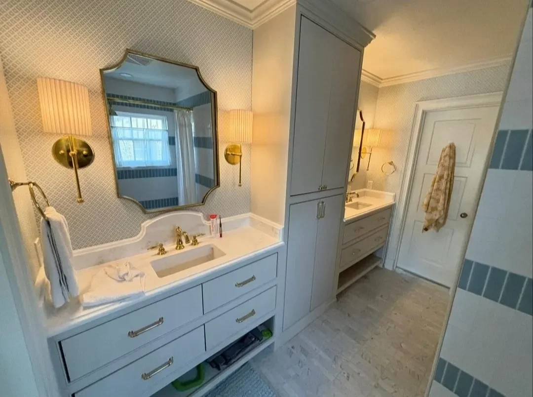 Bathroom with two white vanities, gold fixtures, wall-mounted lamps, large mirror, light-colored tiled walls and floors, and a towel hanging on a hook.