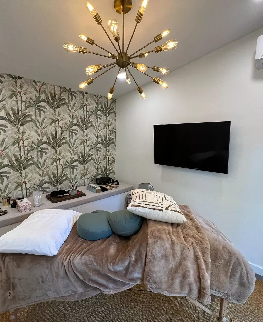 A bedroom with a bed covered in a brown plush blanket, pillows, and decorative cushions. There is a wall-mounted flat-screen TV and a decorative wallpapered wall with a botanical pattern. A modern chandelier with multiple exposed bulbs hangs from the