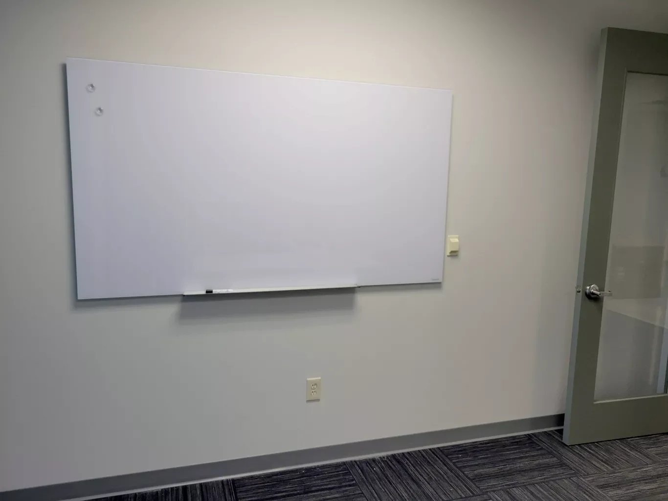 Empty whiteboard on a plain wall in an office or classroom, with a door to the right and electrical outlets below.