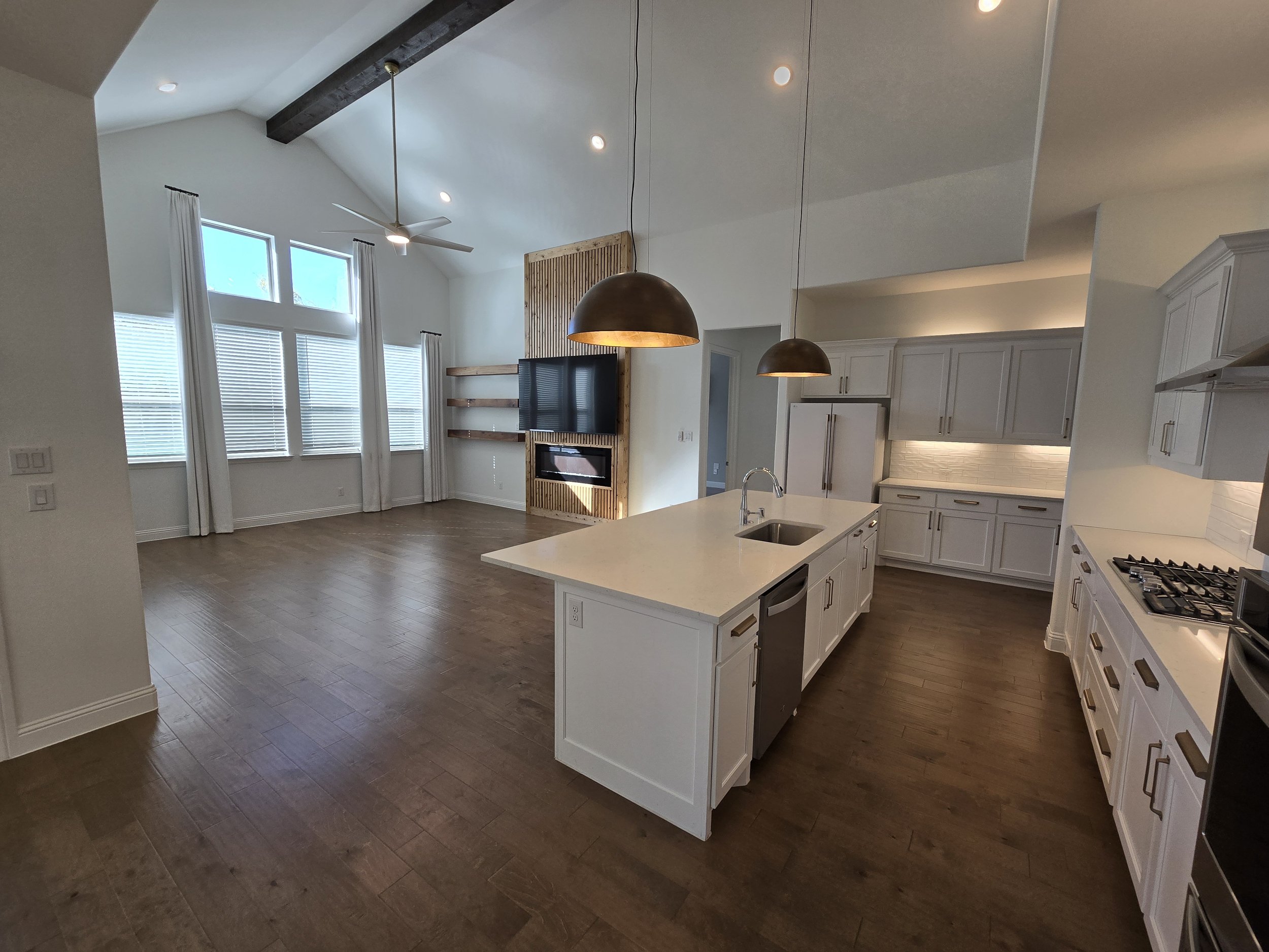 Open-concept kitchen and living room with high ceilings, large windows with white curtains, white cabinets, and dark wood flooring.
