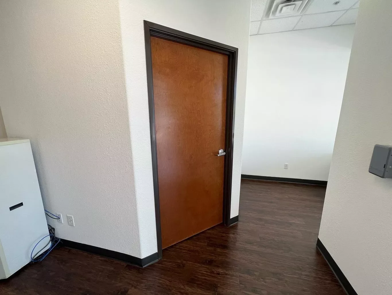 Interior view of an office or commercial space with a closed wooden door, beige walls, dark wood flooring, an electrical panel on the right wall, and a partially visible cabinet on the left.