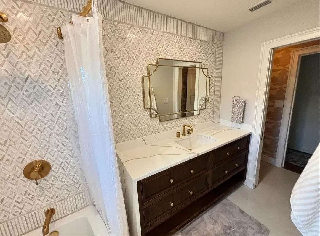 Bathroom with patterned wallpaper, white countertop with double sink and gold fixtures, dark wood vanity, decorative mirror, shower with white curtain, and a small towel hanging on the wall.