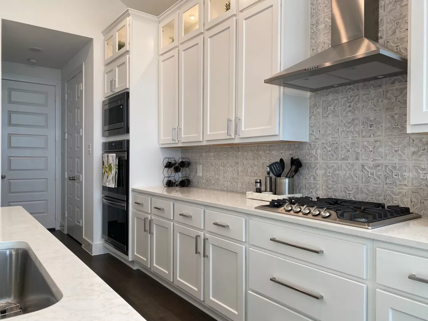 Modern white kitchen with built-in oven and microwave, stainless steel range hood, gray patterned backsplash, white cabinets with silver handles, and dark hardwood floor.
