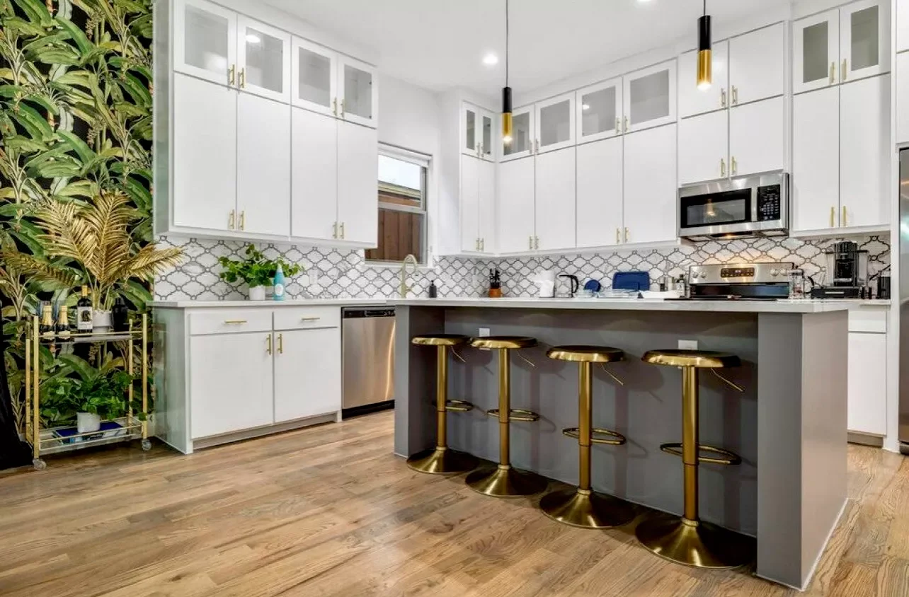 Modern kitchen with white cabinets, a gray island with gold bar stools, hardwood floors, and a patterned white backsplash.