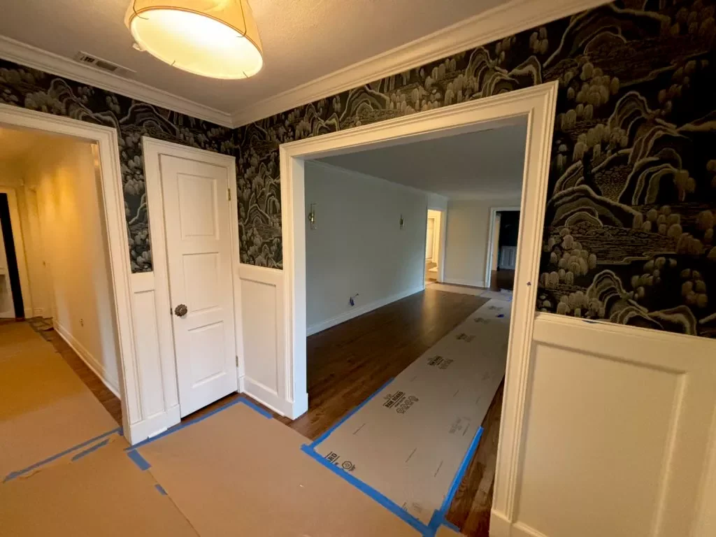 Interior view of a house under renovation, with partially installed flooring, painted walls, and decorative wallpaper. Cardboard covering the new flooring sections, blue painter's tape marking the edges, and doors leading to other rooms.