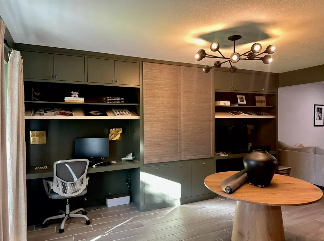 Living room with a built-in green and wood office area, a desk with computer and chair, a round wooden dining table with a black vase and paper roll, a modern ceiling light fixture, and part of a sofa in the background.