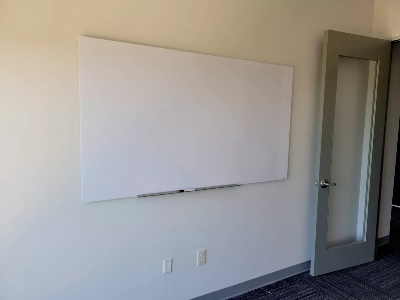Empty whiteboard on a wall in a room with two electrical outlets and a glass-paneled gray door.