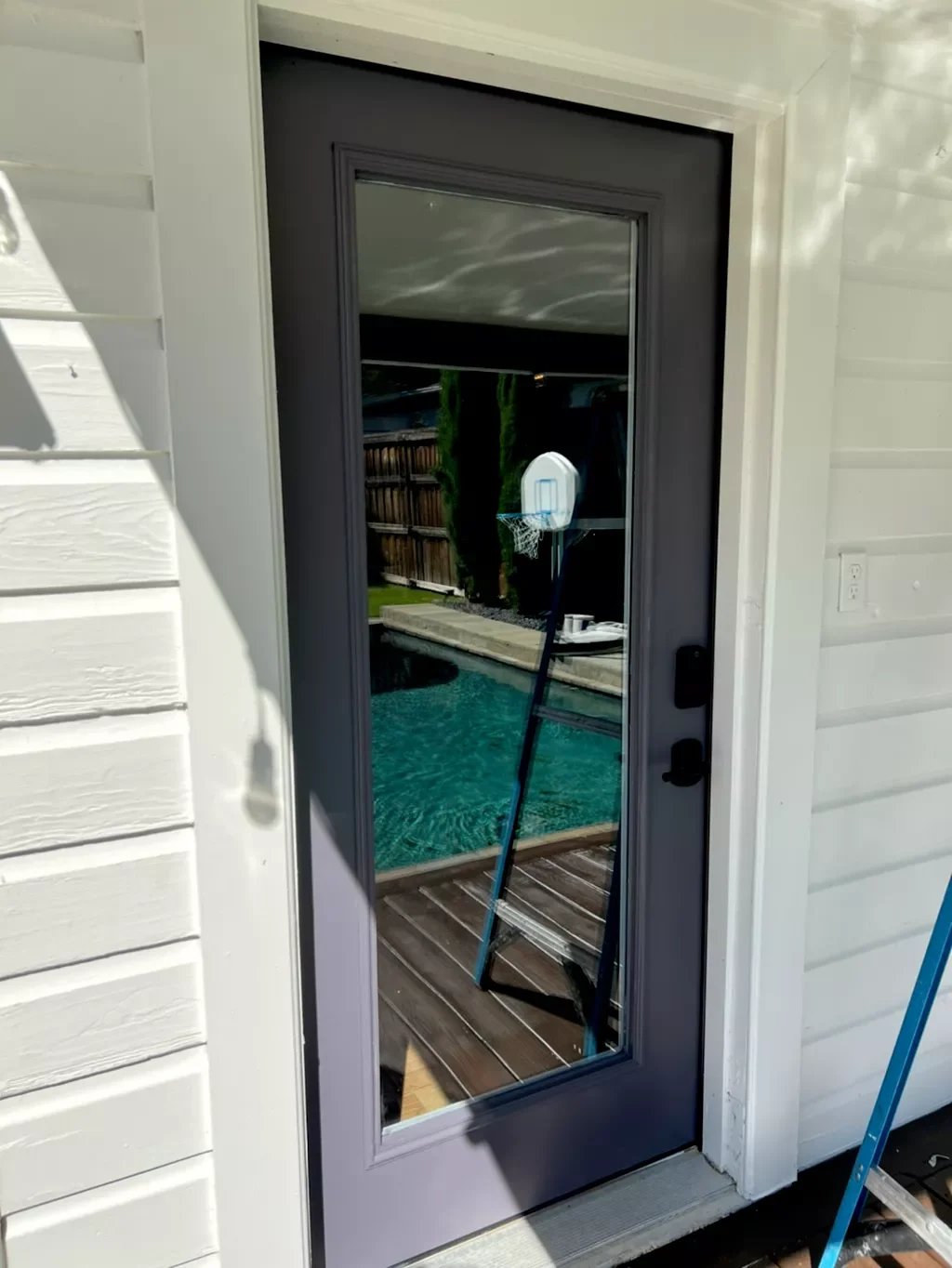 A glass patio door reflects a backyard with a swimming pool, a basketball hoop, and trees.