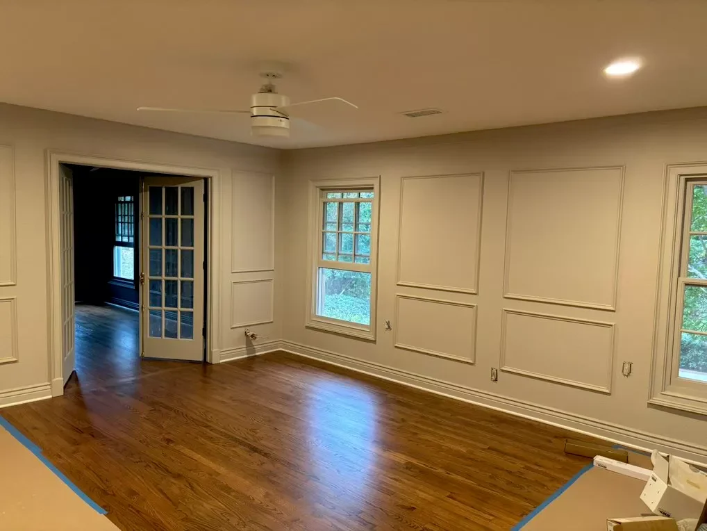 Empty living room with hardwood floors, white walls, decorative wall paneling, a ceiling fan, and large windows showing greenery outside.