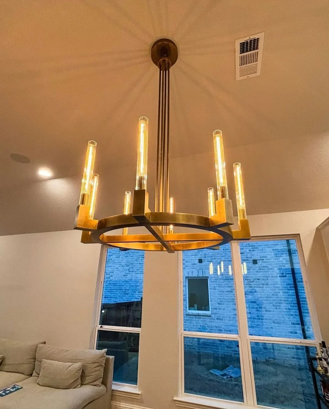 Modern chandelier with exposed filament bulbs hanging from the ceiling in a living room, with large window showing a brick building outside.