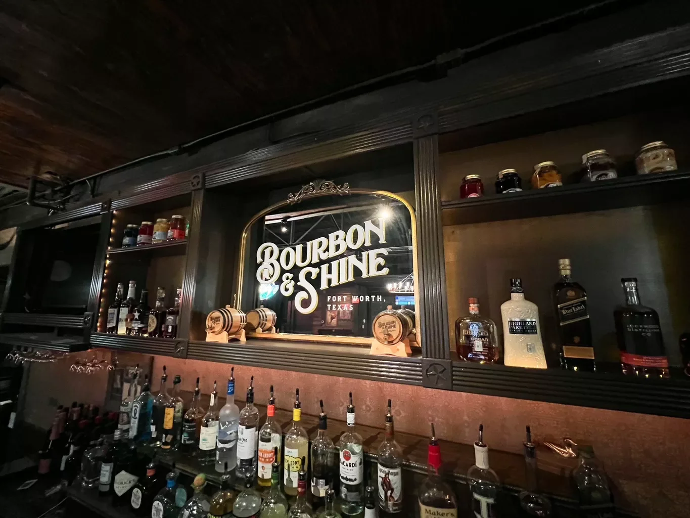 Bar area with a sign that says 'Bourbon & Shine' in Fort Worth, Texas. Bottles of liquor on shelves and a variety of alcohol bottles on the bar counter.