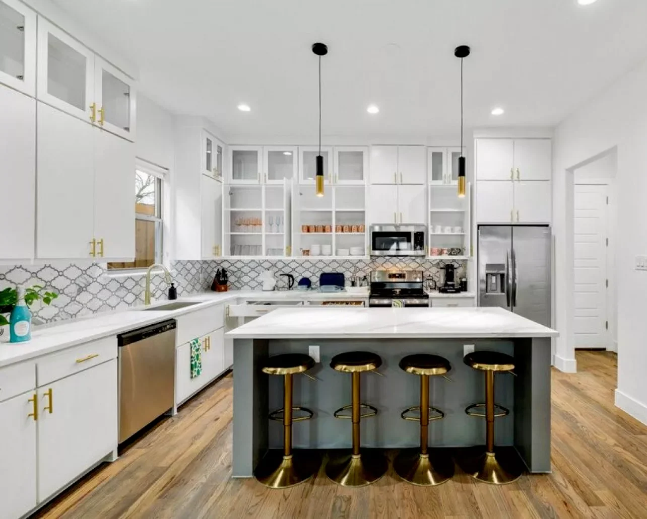 Modern white kitchen with gold hardware, wooden floor, and central island with four black and gold stools, pendant lights, stainless steel appliances, open shelving, and a window over the sink.