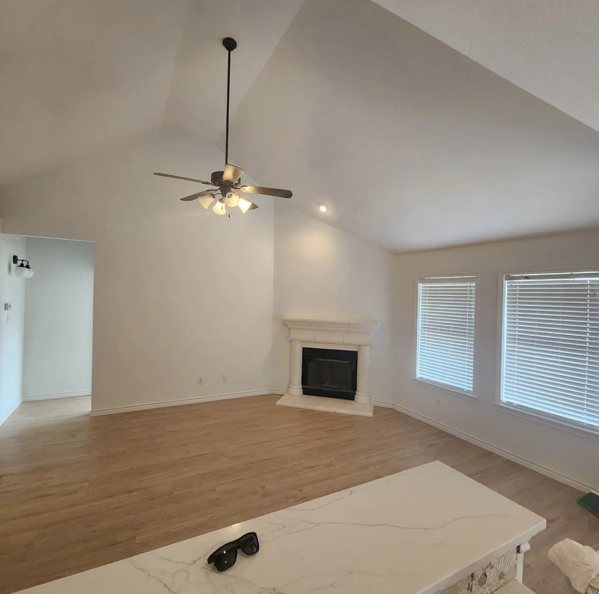 Empty living room with hardwood floors, white walls, a ceiling fan with lights, a fireplace, and large windows with blinds.