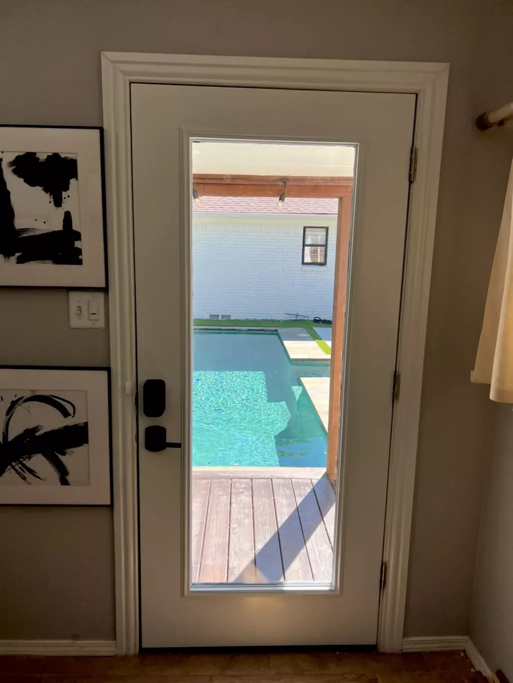 View through a glass door opening to a backyard pool area with a wooden deck and a white house in the background.