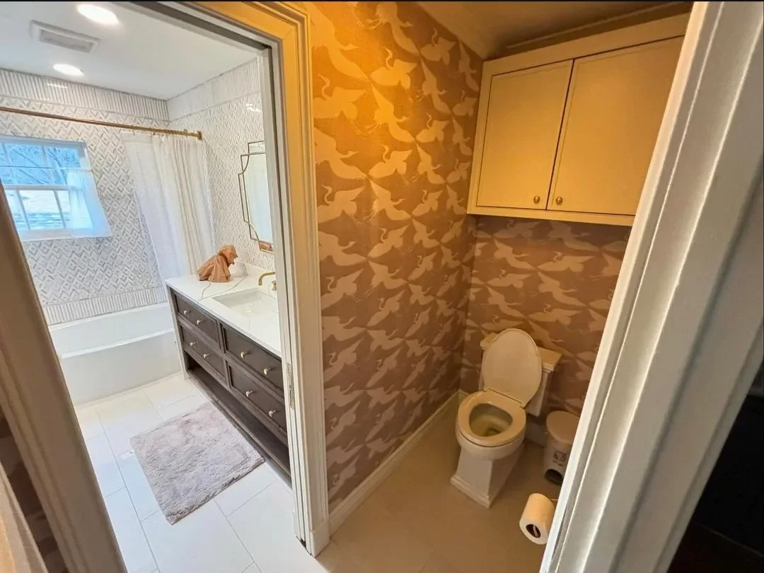 View of a bathroom with a toilet and a small cabinet, the main bathroom area on the left has a bathtub, sink, mirror, window, and shower curtain.