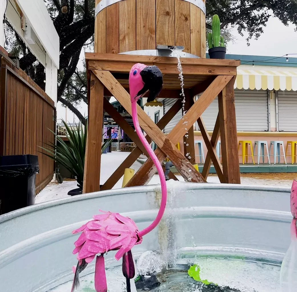Pink flamingo-shaped fountain with water flowing from its neck into a round container, outdoor setting with wooden structure, potted cactus, and colorful bar stools.