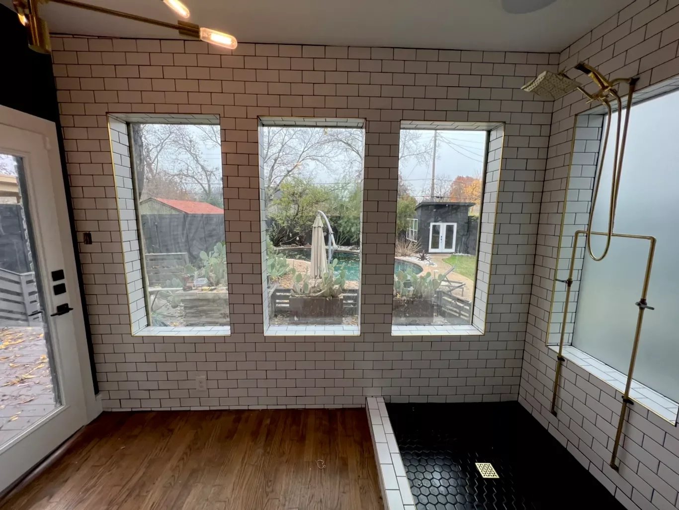 Bathroom with white subway tile walls, three large windows looking out to a backyard with plants, trees, and a pool, wooden floor, and a black shower area with gold fixtures.