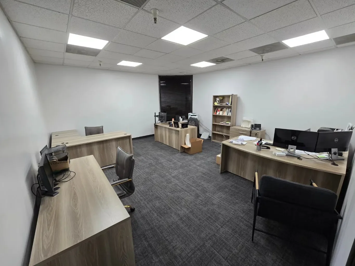 Empty office room with four desks, some computers, chairs, bookshelves, and office supplies.