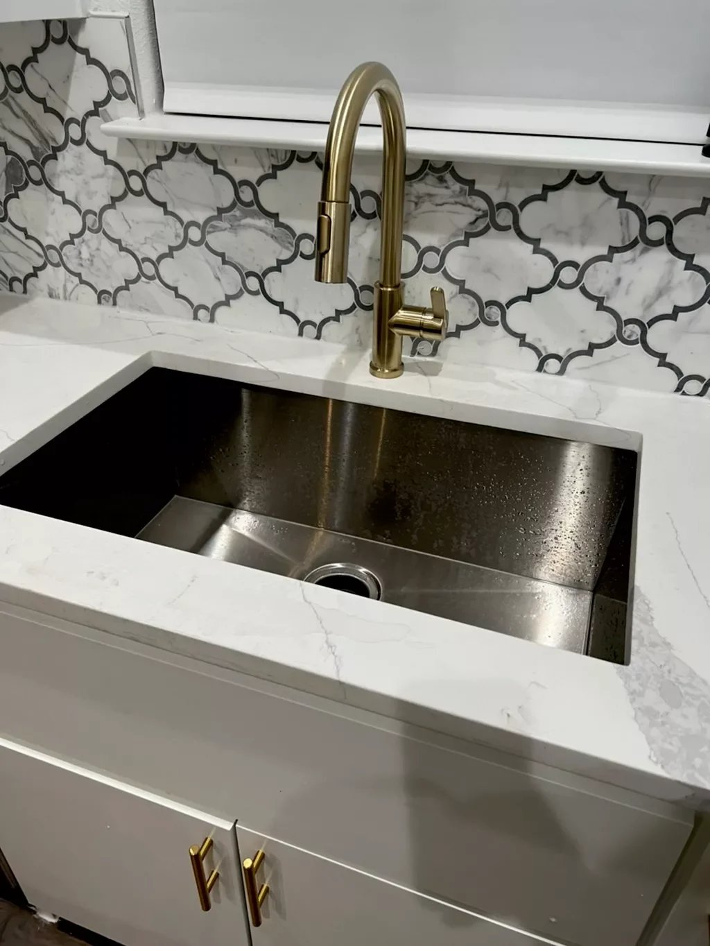 Kitchen sink with a gold faucet, white marble countertop, and a decorative black and white tile backsplash.