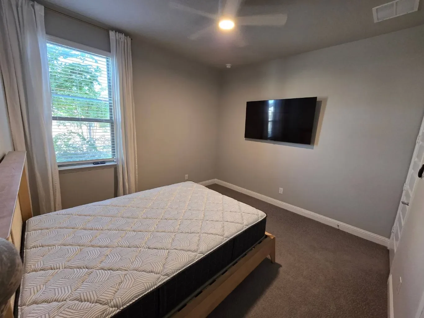 A bedroom with a window, a twin bed with a patterned mattress, a mounted flat-screen TV on the wall, and a ceiling light.