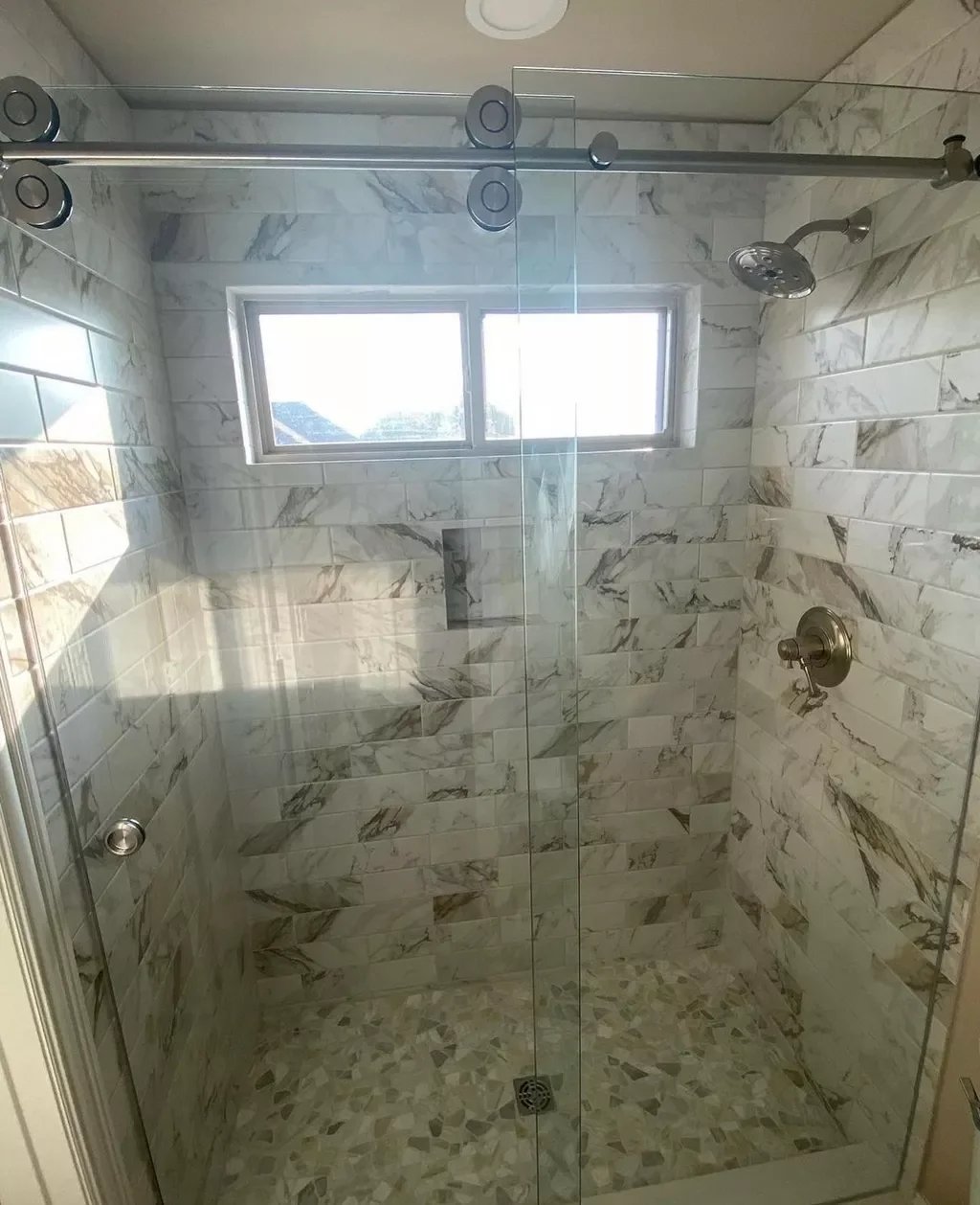 A modern walk-in shower with marble tiles on the walls and floor, a small window on the back wall, a ceiling-mounted showerhead, and a glass enclosure.