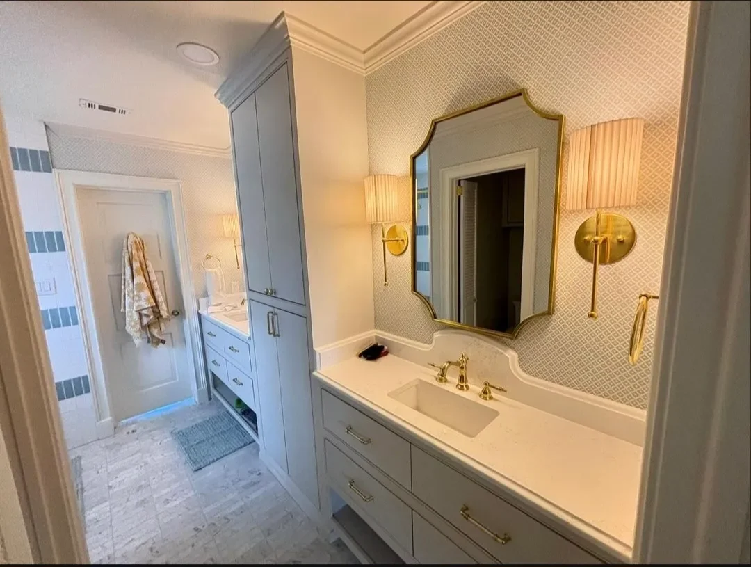 Bathroom with a vanity featuring a mirror, gold fixtures, and a white countertop. Wall lamps with pink shades provide lighting. Towels hang on a door, and an additional cabinet is seen beside the vanity.