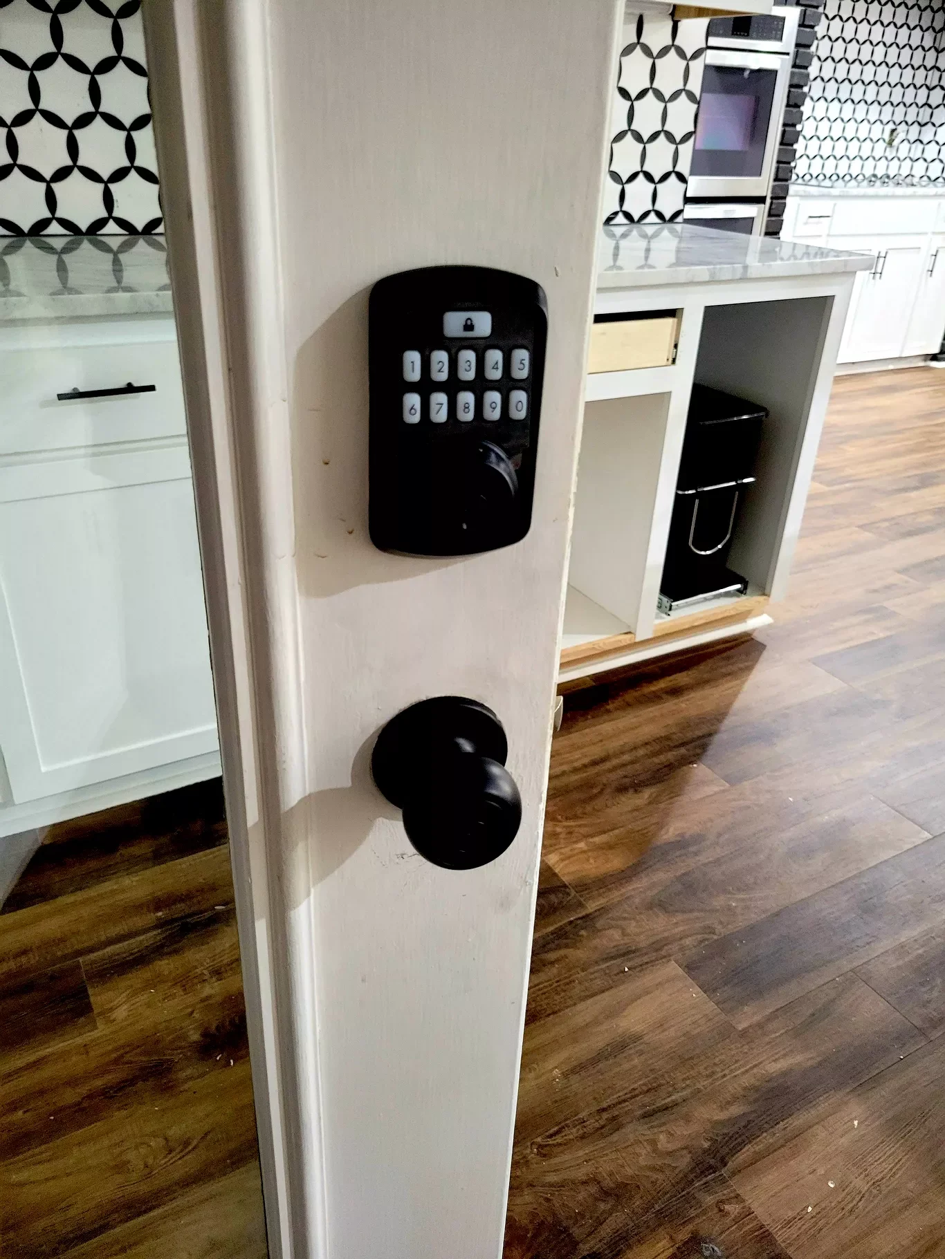 Door with a keypad lock and a doorknob, beside a kitchen with wooden flooring, white cabinets, and black and white patterned backsplash.