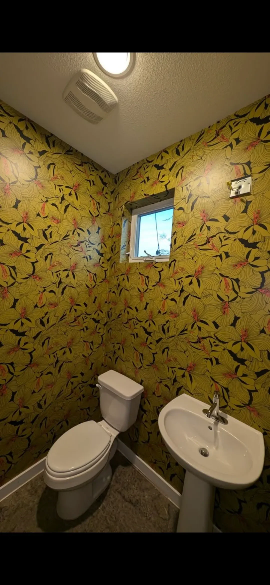 Small bathroom with floral yellow wallpaper, white toilet, white sink, small window, ceiling light, and wall vent.