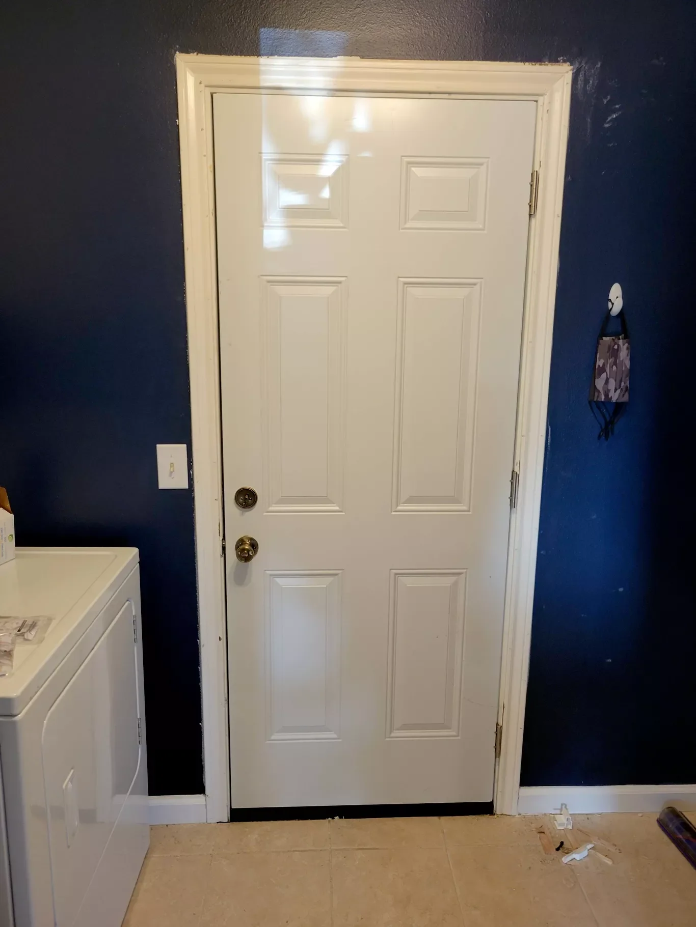 A white door with six raised panels, a spherical doorknob, and a keyed lock, set in a dark blue wall, with a white washing machine to the left and a purple face mask hanging on the right wall.