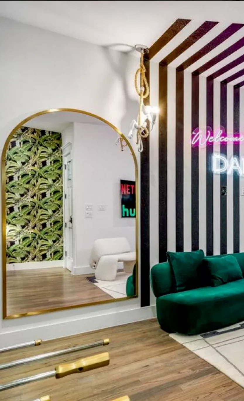 Interior of a modern living room with a large gold-framed mirror, a green velvet couch, striped black and white walls, a neon 'Welcome' sign, and a patterned wallpapered wall with tropical leaves in the background.