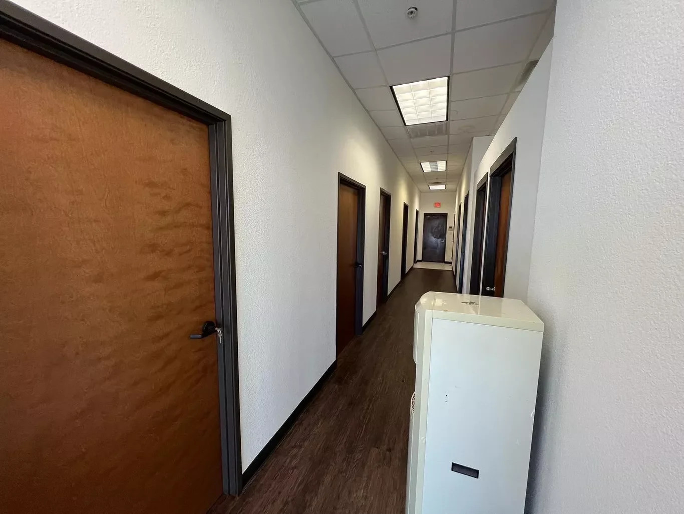 Empty hallway with wooden doors, white walls, and raining light fixtures on ceiling. Water fountain on right side.