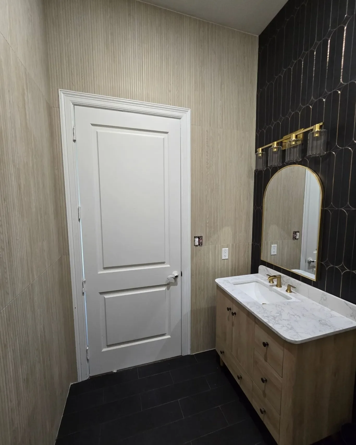 Bathroom with a white wooden door, a wall-mounted mirror with a gold frame, black tiled wall behind the mirror, a marble countertop with a gold faucet, and a wooden vanity cabinet underneath.