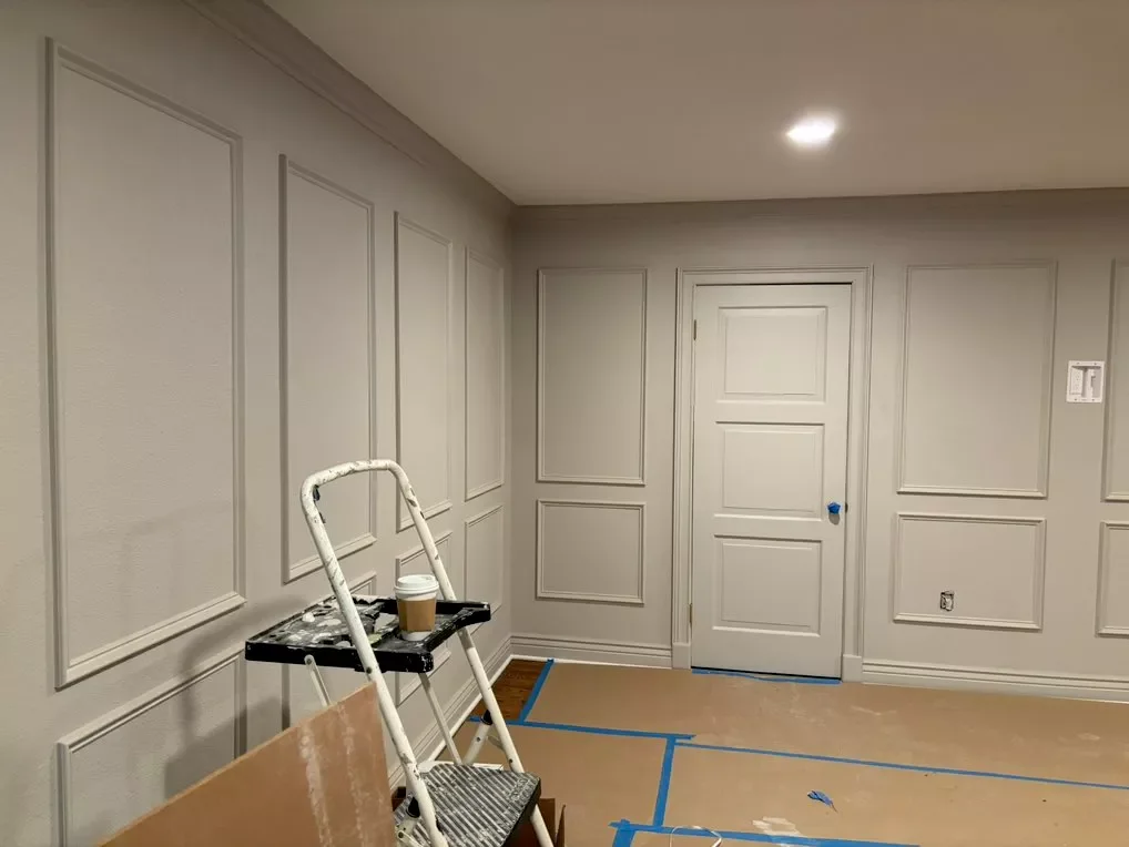Interior of a room under renovation with light gray paneled walls, a closed white door, and painter's tape on the floor. A small stepladder with a coffee cup and paint supplies is in the corner.