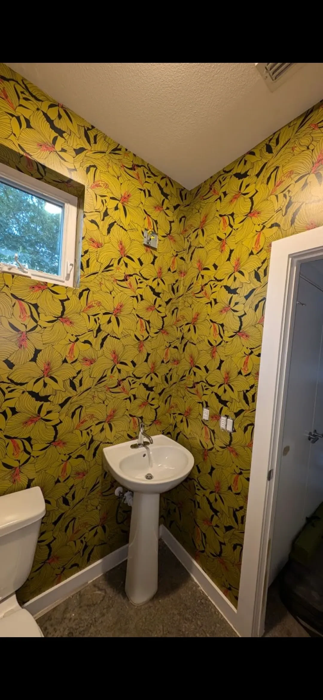A small bathroom with yellow floral wallpaper, a white pedestal sink, a toilet, a small window, and an air conditioning vent on the ceiling.