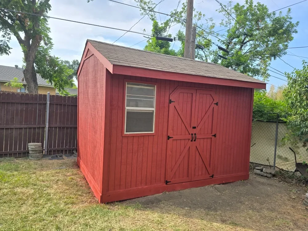 Red wooden shed with double doors and a small window, situated in a backyard with a fenced yard and trees.