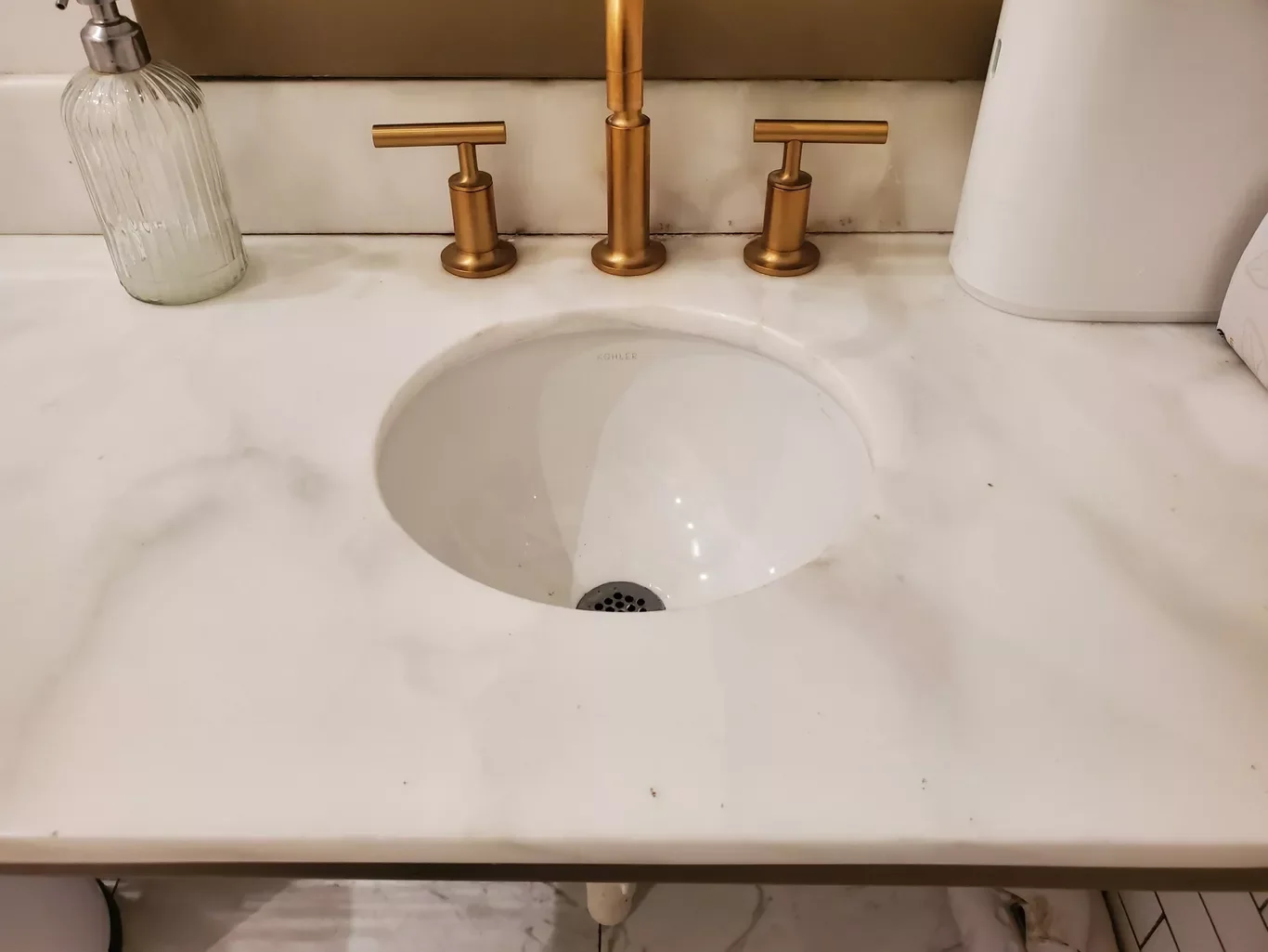 Bathroom sink with gold-colored faucet and handles, a clear soap dispenser on the left, and a white tissue box on the right, installed on a white marble countertop.