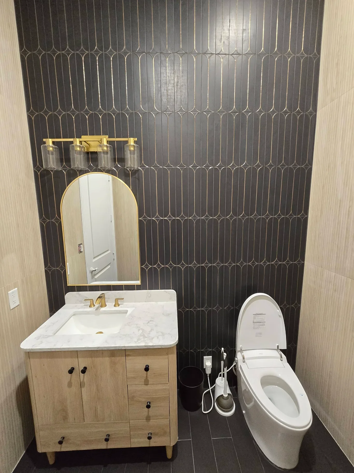 A modern bathroom with a white toilet, a wooden vanity with a marble countertop and gold faucet, a vertical mirror with a gold frame, a black wall-mounted light fixture, and black tiled walls.