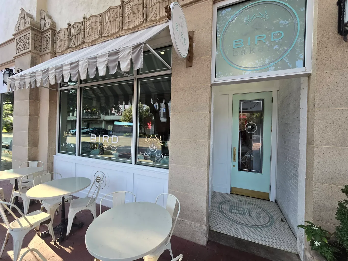 Exterior of Bird Bakery with outdoor seating, cream-colored tables and chairs, a large window with the bakery's name, and a door with a nearby sign above it.