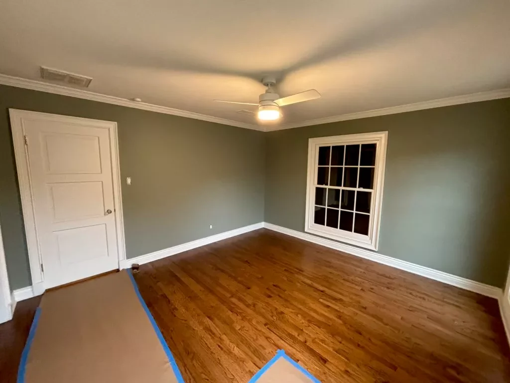 Empty room with hardwood floor, green walls, a white door, a large window with white trim, and a ceiling fan with light.