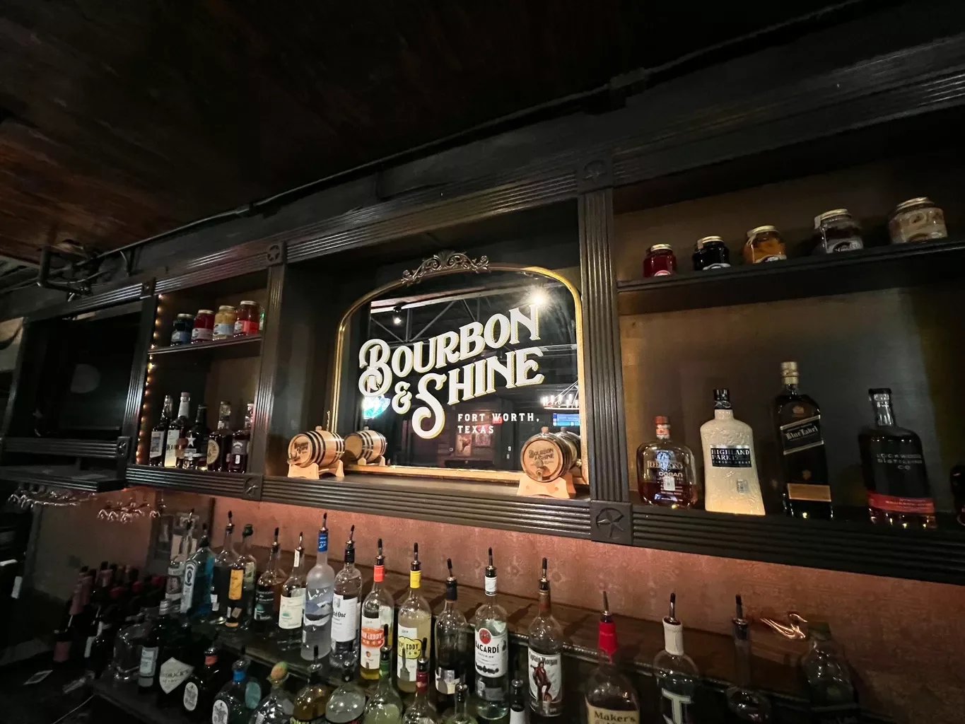 Bar area with a sign that says 'Bourbon & Shine' in Fort Worth, Texas. Bottles of liquor on shelves and a variety of alcohol bottles on the bar counter.