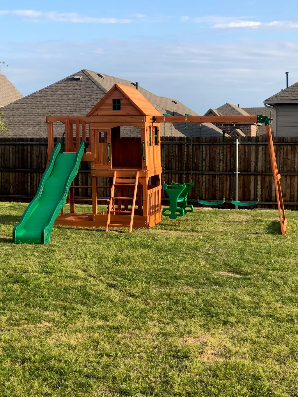 A backyard playground featuring a wooden playset with a green slide, a small house with a peaked roof, a baby swing, and a regular swing, enclosed by a wooden fence, with neighboring houses in the background under a partly cloudy sky.