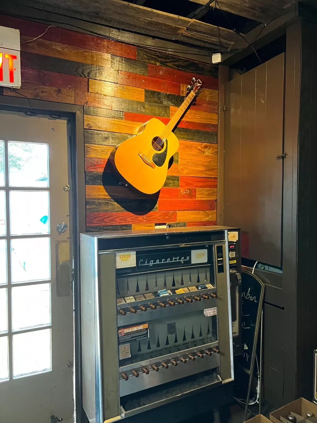 A wooden wall with a guitar mounted on it, next to a cigarette vending machine in a rustic-style interior.