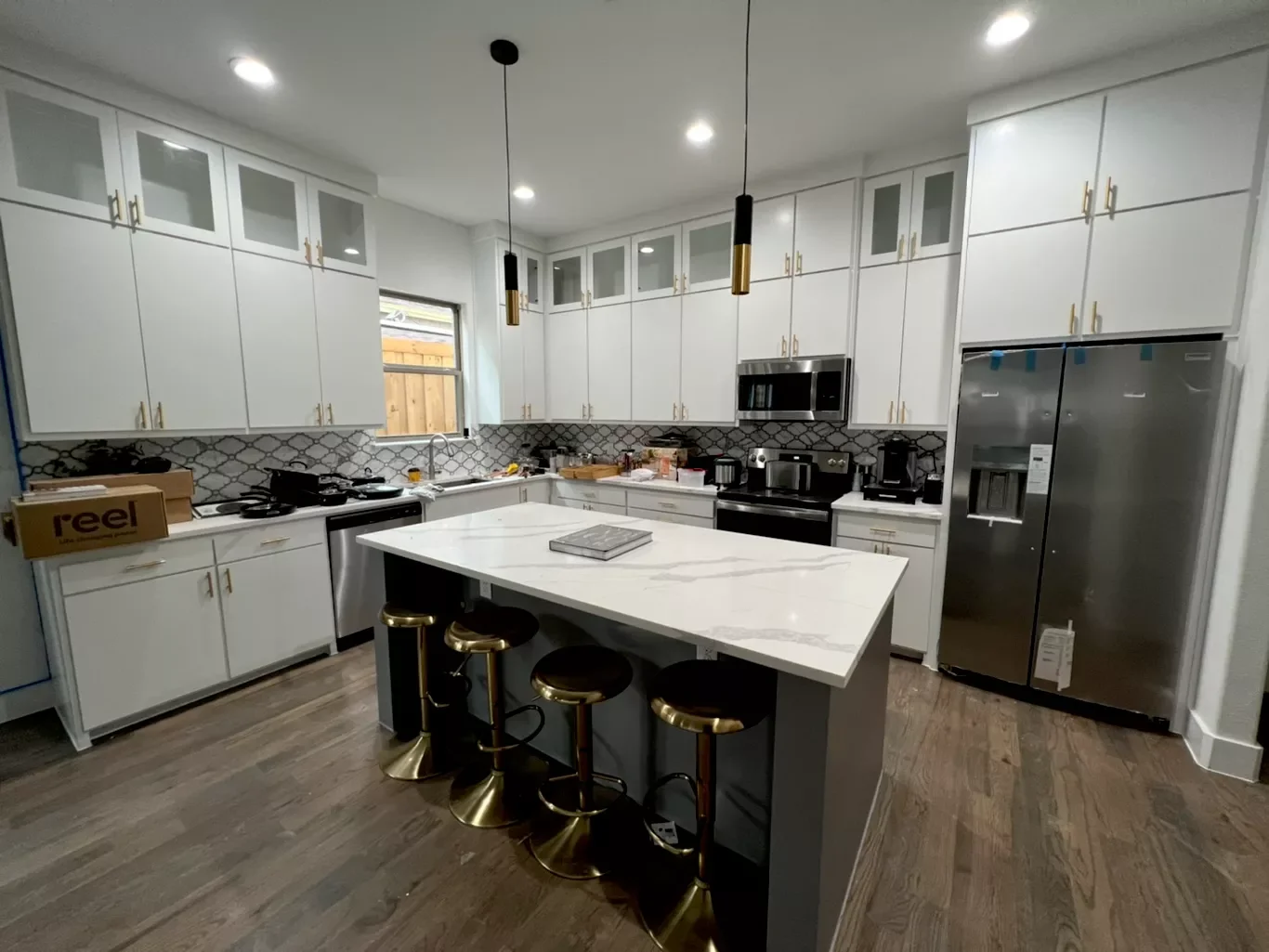 Modern kitchen with white cabinets, black and gold pendant lights, stainless steel appliances, marble countertop island, and wood flooring.