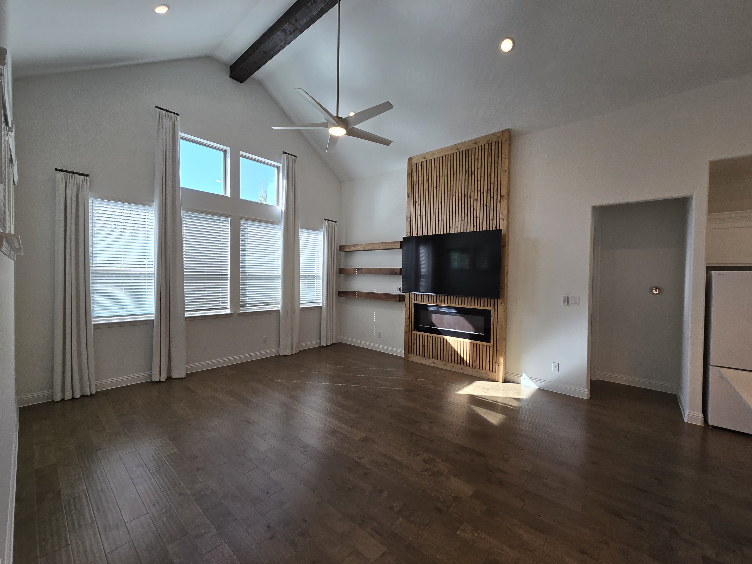 Empty living room with large windows, white walls, hardwood flooring, and a ceiling fan. There is a mounted black TV on a wooden accent wall with a built-in fireplace below, and two wooden shelves to the right of the TV.