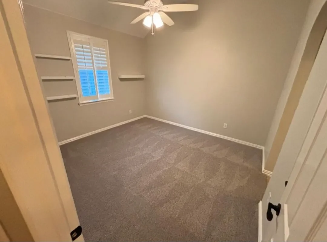 Empty bedroom with beige walls, a ceiling fan, a window with plantation shutters, and built-in wall shelves.