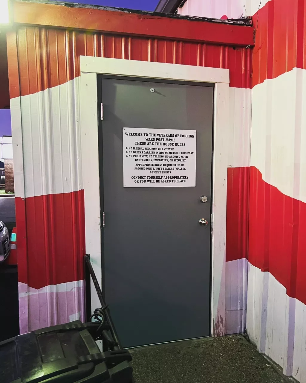 A grey door with a sign, set in a building with red, white, and pink painted walls, and a nearby black trash can.