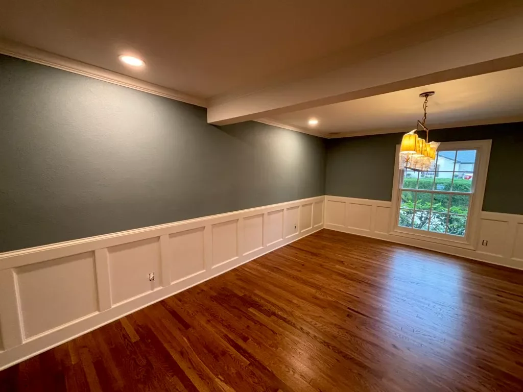 Empty dining room with wooden floors, green walls, white wainscoting, and a window with a view of greenery outside. There is a modern chandelier hanging from the ceiling.