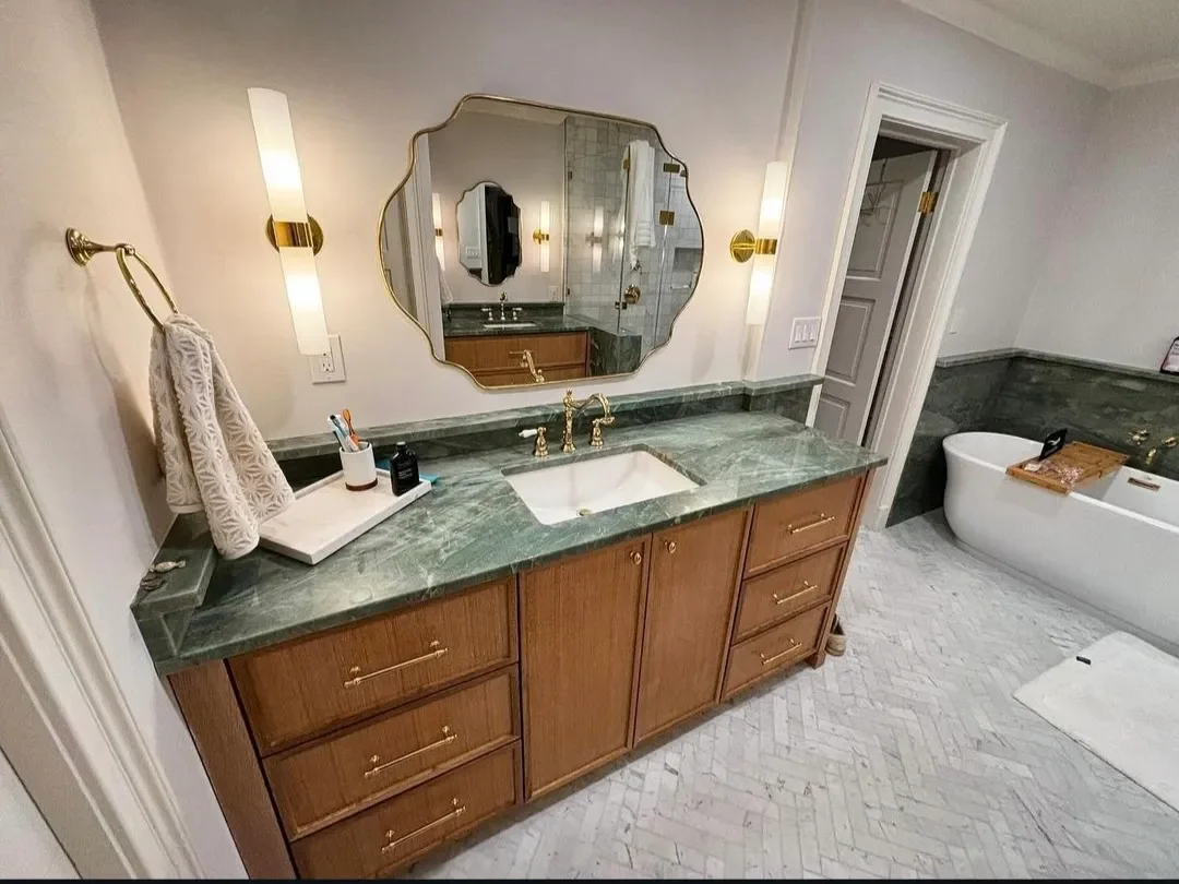A bathroom with a green marble countertop, a rectangular sink, and a large ornate mirror. There are two wall-mounted lights flanking the mirror, a towel on a ring on the left, and a wooden cabinet under the sink. In the background, there is a bathtub