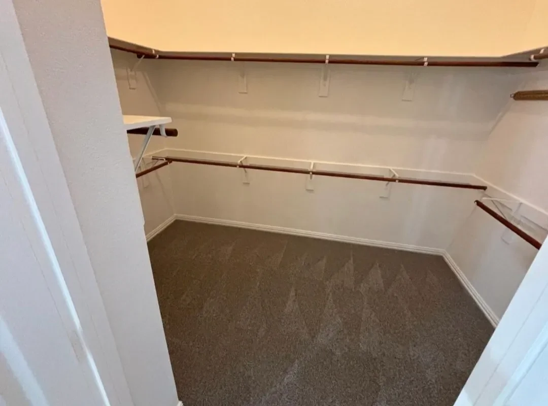Empty walk-in closet with beige carpet, white walls, and multiple wooden and metal clothing rods on three sides.